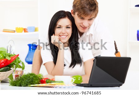 Beautiful young couple cooking looking at laptop screen with receipt in the kitchen, happy smile