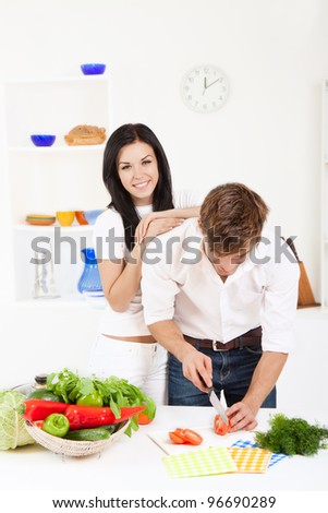 portrait of young lovely couple cooking slicing tomato in their kitchen, woman embrace, hug happy smile looking at camera