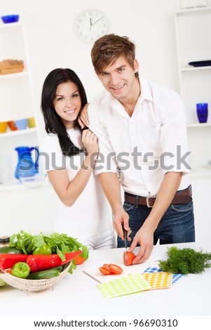 portrait of young lovely couple cooking slicing tomato in their kitchen, looking at camera woman embrace, hug happy smile