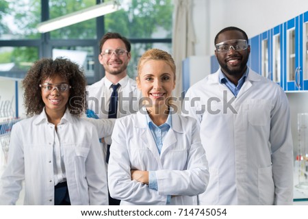 Smiling Group Of Scientists In Modern Laboratory With Female Leader, Mix Race Team Of Scientific Researchers In Lab Wearing White Coats And Protective Glasses