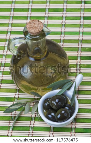 A bottle of olive oil  and a china spoon with olives on a bamboo mat