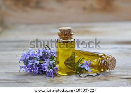 Rosemary essential oil in a small glass vial and plant with flowers on a wooden background