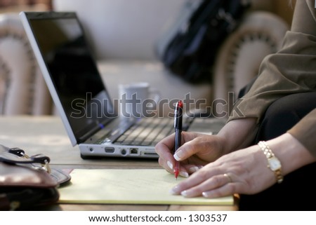 Businesswoman Writing Notes next to her Laptop Computer in a Hotel Atrium (shallow focus point on hand with pen).