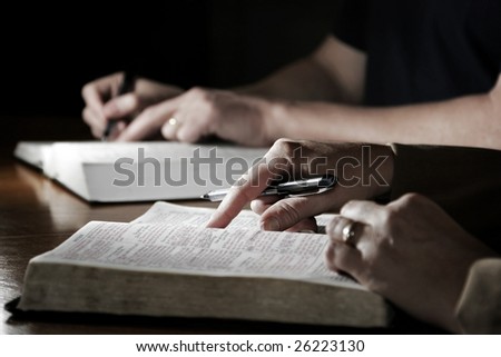 A man and woman (or married couple) study their Holy Bibles together at a table - slight color effect, shallow focus point on foreground woman\'s hand.