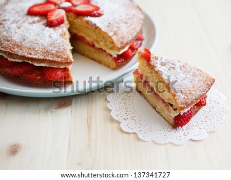 Victoria sponge cake with straberries, jam and whipped cream with a cut out piece on a wooden table
