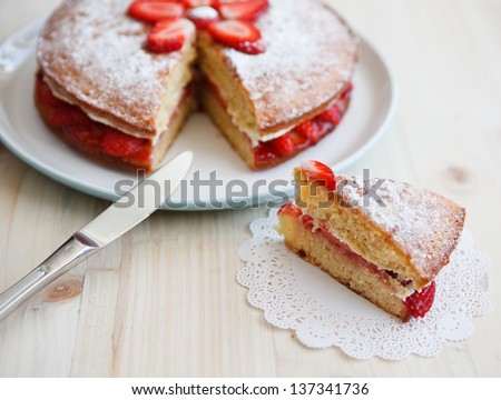 Victoria sponge cake with strawberries, jam and whipped cream with a cut out piece and a knife on a wooden table