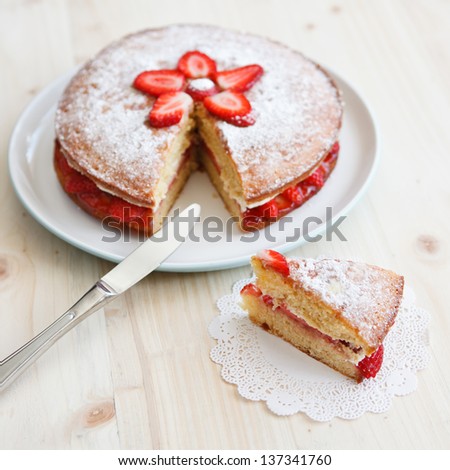 Victoria sponge cake with strawberries, jam and whipped cream with a cut out piece and a knife on a wooden table, square