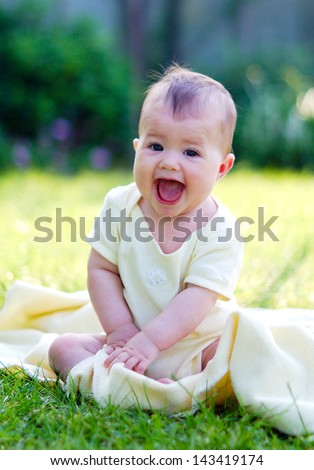 A happy laughing baby in a yellow vest sitting on the blanket in the park