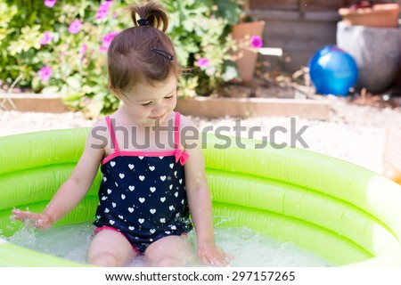 A little toddler girl in a black swimsuit playing in a green paddling pool in the garden