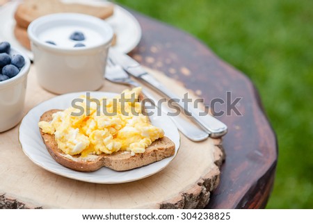 Breakfast in the garden: scrambled eggs on toast and yogurt with berries on the dark wooden table, selective focus
