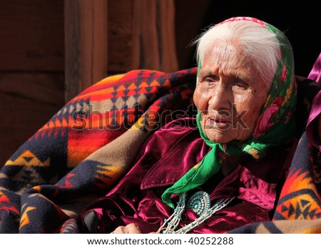 An elderly Native American woman sits among blankets. She is head and shoulders viewable and looking away from the camera. Horizontally framed shot.