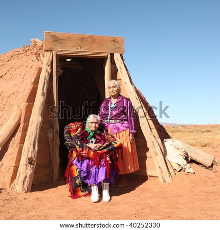 Elderly Native American women pose outside a tribal abode. They are full length viewable and looking at the camera. Squarely framed shot.