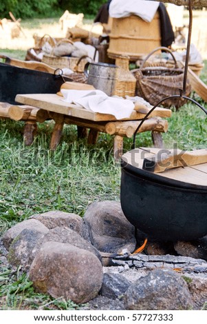 A stone fire pit, with a cast iron pot outdoors. A small preparation table and other cooking utensils are placed nearby in this open-air medieval style kitchen