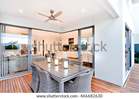 Outdoor patio area with table set up beside an entrance to inside of a modern house with a kitchen, there are candles on the table under the fan