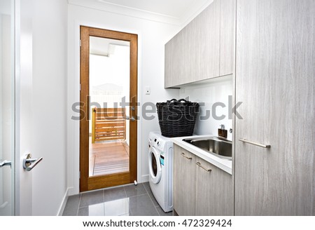 Modern laundry room with grey cupboards on the wall near a wooden door with a glass panel closed, there is a black basket made in rattan on the washing machine