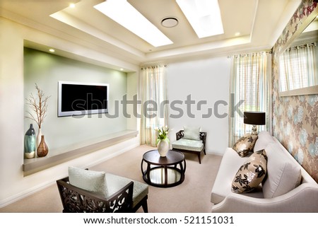 This luxury living room has a long white sofa with pillows  close to flower paint decoration on the wall, There are two wooden chairs with pillows near a round table with ornamental items.