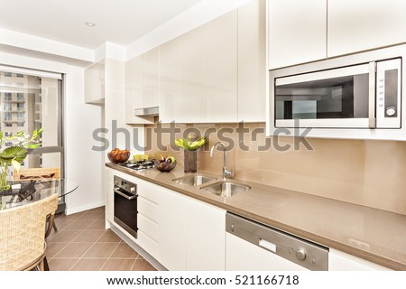 Kitchen with luxurious tools and white walls, luxury living room including glass table also chairs, oven and gas cooker have attached to the pantry cupboard, ceramics with fruits near the cookers.