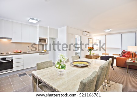 Luxury kitchen with modern table and sofa, wash basin also gas cooker have attached to the wall, flower pot near ceramic plate on  dining table, chairs are cushioned in white color.