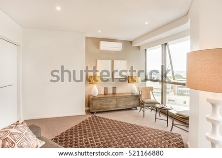 Living room with city view near glass windows, sofa set including pillows, lamps on the wooden table, walls are white color, floor carpet has some designs, daytime scene also perfect lightning.