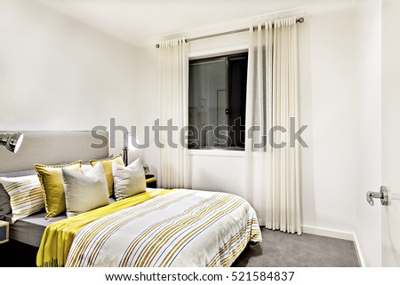 Classic bedroom of a modern house with table lamps on next to pillows and bed with duvets, there is a black glass window covered with a curtain