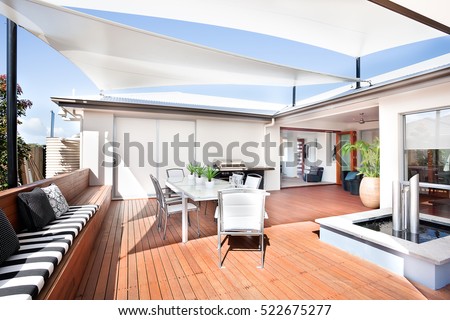 This patio area has a wooden floor and a long bench attached to it, there are some small mattresses and pillows on it. A white table and chairs are under the creative roof design,