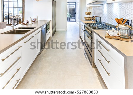 Tile floor with a hallway through the kitchen with ovens, stoves, taps with sink fixed to the counter, there are utensils on the cupboards
