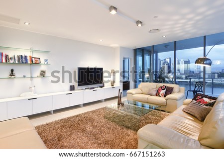 Living room with books near television, comfortable furniture and designs, walls are white color, chairs around the tables, inside rooms of a apartment.