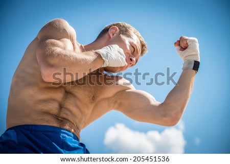 Athletic guy in blue shorts and boxing gloves fulfills uppercut punch, exercising in nature