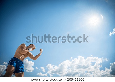 Athletic guy in blue shorts and boxing gloves fulfills uppercut punch, exercising in nature