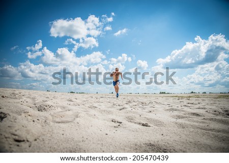 Athletic guy in blue shorts and sunglasses running along the golden sand and blue sky