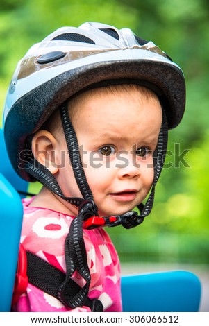 Little girl dressed in bicycle helmet and sits on a bicycle seat for children. Emotions: curiosity