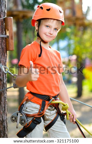Cute boy shows thumb up with climbing equipment in an adventure park