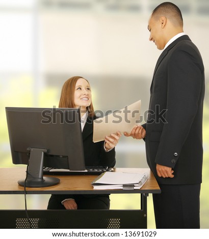a business man hand over a folder to woman sitting with computer. both showing smiling and happy expression. concept for team work, business related, or business deal