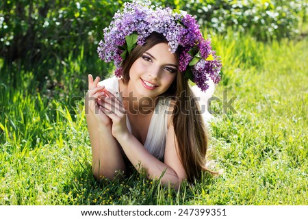 Beautiful smiling teen girl is lying on grass with fashion makeup is wearing beautiful wreath of lilac flowers
