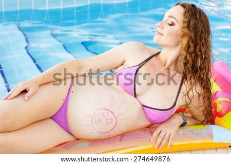 Beautiful pregnant woman is wearing swimsuit is lying and resting on colorful mattress near swimming pool with blue water, vacations