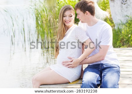 Happy pregnant woman with her husband are sitting on wooden bridge near blue river, summer time, pregnancy girl