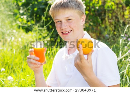 Portrait of cute smiling teenager boy with freckles on his face is holding glass with orange juice and fruits, summer time