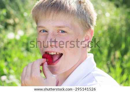 Portrait of cute smiling boy with freckles on his face is sitting on green grass with red strawberry