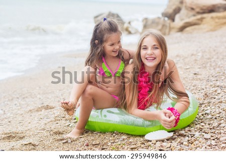 Two pretty smiling sisters are wearing colorful hawaiian flowers and swimsuits sitting on pink rubber ring at the beach, summer time