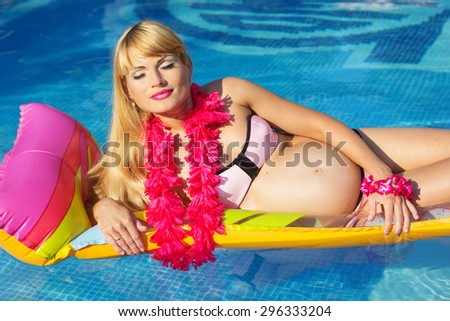Beautiful pregnant woman is wearing pink hawaiian flowers lying on colorful mattress in blue water of swimming pool