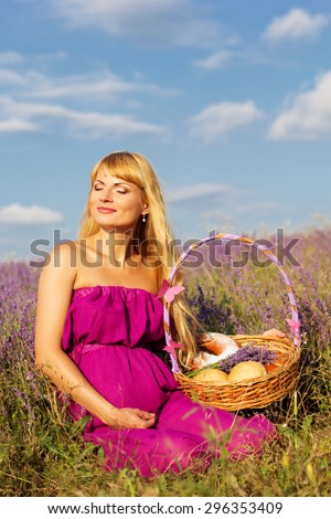 Beautiful pregnant blonde woman is wearing purple dress is holding basket with purple ribbon and sweet buns and rolls