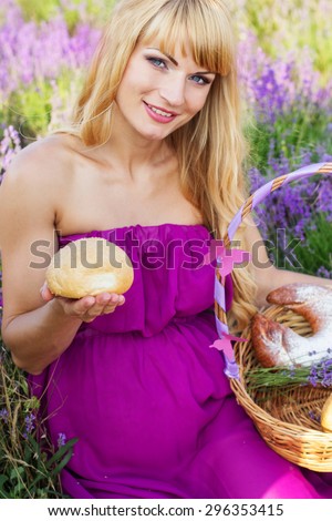 Beautiful pregnant blonde woman is wearing purple dress is holding basket with purple ribbon and sweet fresh buns and rolls