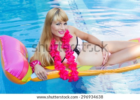 Beautiful pregnant woman is wearing pink hawaiian flowers lying on colorful mattress in blue water of swimming pool, summer vacations