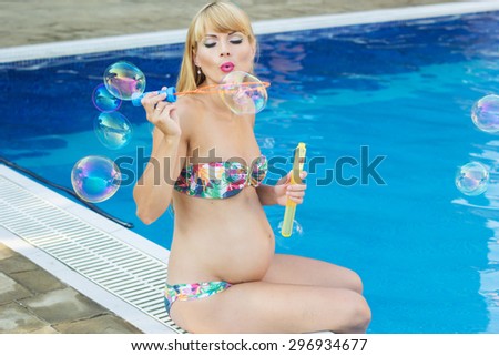 Beautiful pregnant woman is making soap bubbles near swimming pool with blue water, vacations