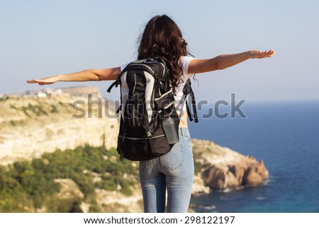 Back view of young pretty slim woman standing with backpack on cliff\'s edge and looking into a wide valley