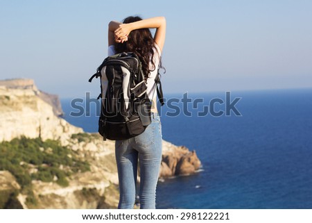 Back view of young pretty slim woman standing with backpack on cliff\'s edge and looking into ocean