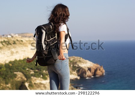Back view of young pretty woman standing with backpack on cliff\'s edge and looking into a wide valley