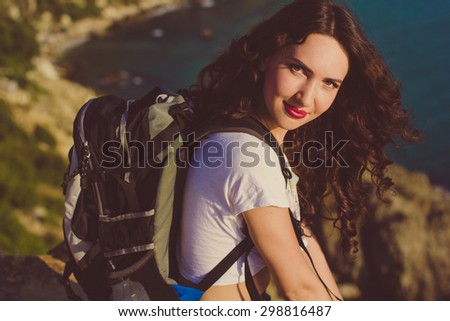 Pretty smiling traveler girl backpacker is drinking water on rock peak over sea view with backpack, summer vacations