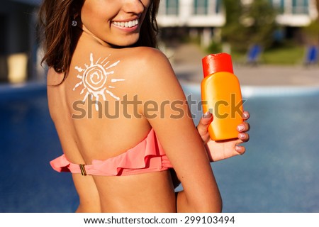 Sexy smiling girl with sun shape drawing from sunscreen lotion on her shoulder is resting near swimming pool with blue water, summer time