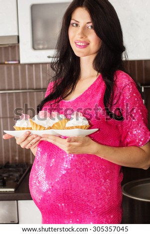 Pregnant happy woman is wearing pink dress and staying in kitchen with sweet cakes